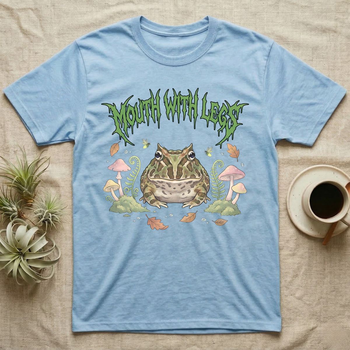 Pacman Frog Feral T-Shirt in Light Blue — Round Pacman frog in a pastel forest-floor scene under a spiky leaf-green metal wordmark reading MOUTH WITH LEGS.