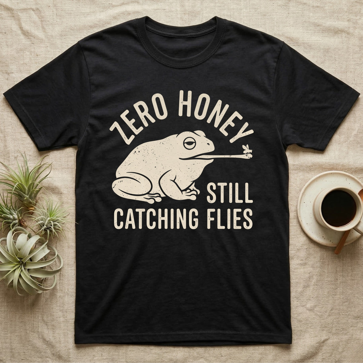 Tree Frog Proverb T-Shirt in Black — Worn cream print of a dumpy White's tree frog catching a fly with its tongue under the words Zero Honey, Still Catching Flies