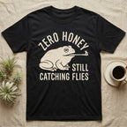 Tree Frog Proverb T-Shirt in Black — Worn cream print of a dumpy White's tree frog catching a fly with its tongue under the words Zero Honey, Still Catching Flies