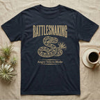 Coiled corn snake mid-tail-vibration in warm tan ink engraving beneath an arched wordmark reading RATTLESNAKING and the subtitle Angry Velcro Mode. — shown on a Navy tee.