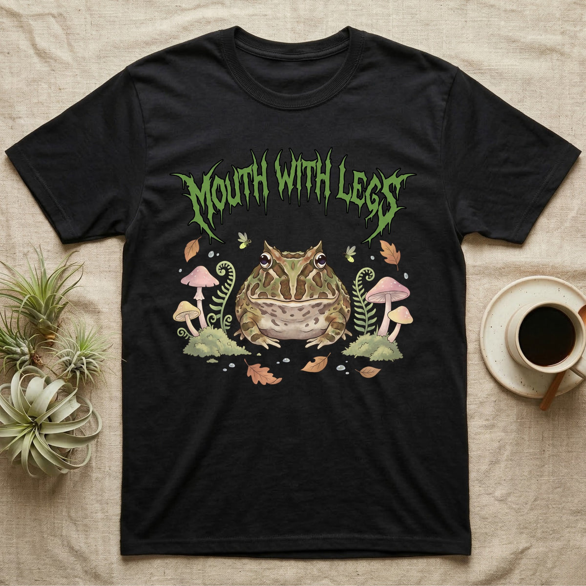 Pacman Frog Feral T-Shirt in Black — Round Pacman frog in a pastel forest-floor scene under a spiky leaf-green metal wordmark reading MOUTH WITH LEGS.