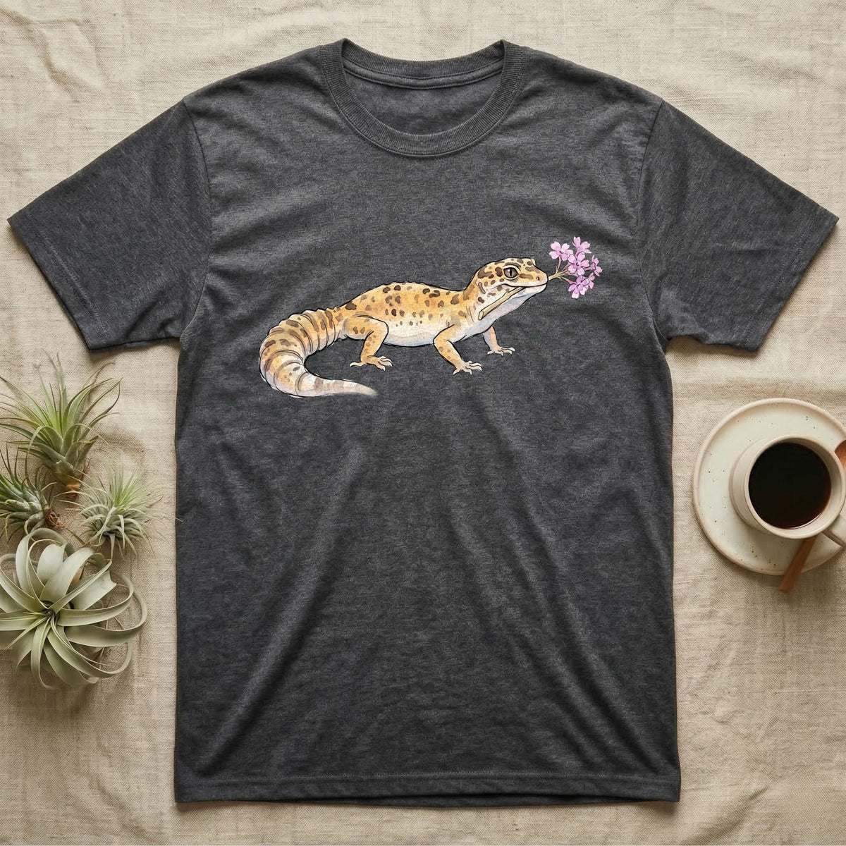 Leopard Gecko Blossom T-Shirt in Dark Heather — Watercolor illustration of a sandy-gold spotted leopard gecko holding a sand verbena blossom sprig, on a dark grey t-shirt.