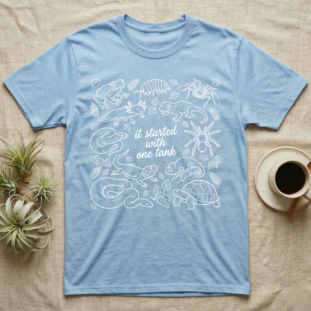 Terrarium Keeper One Tank T-Shirt in Light Blue — White outline illustration of eleven terrarium species — bearded dragon, crested gecko, ball python, axolotl, tortoise, tarantula, isopod, whip spider, chameleon, tree frog and hognose — with pothos and fern leaves, ringed around the script line it started with one tank.