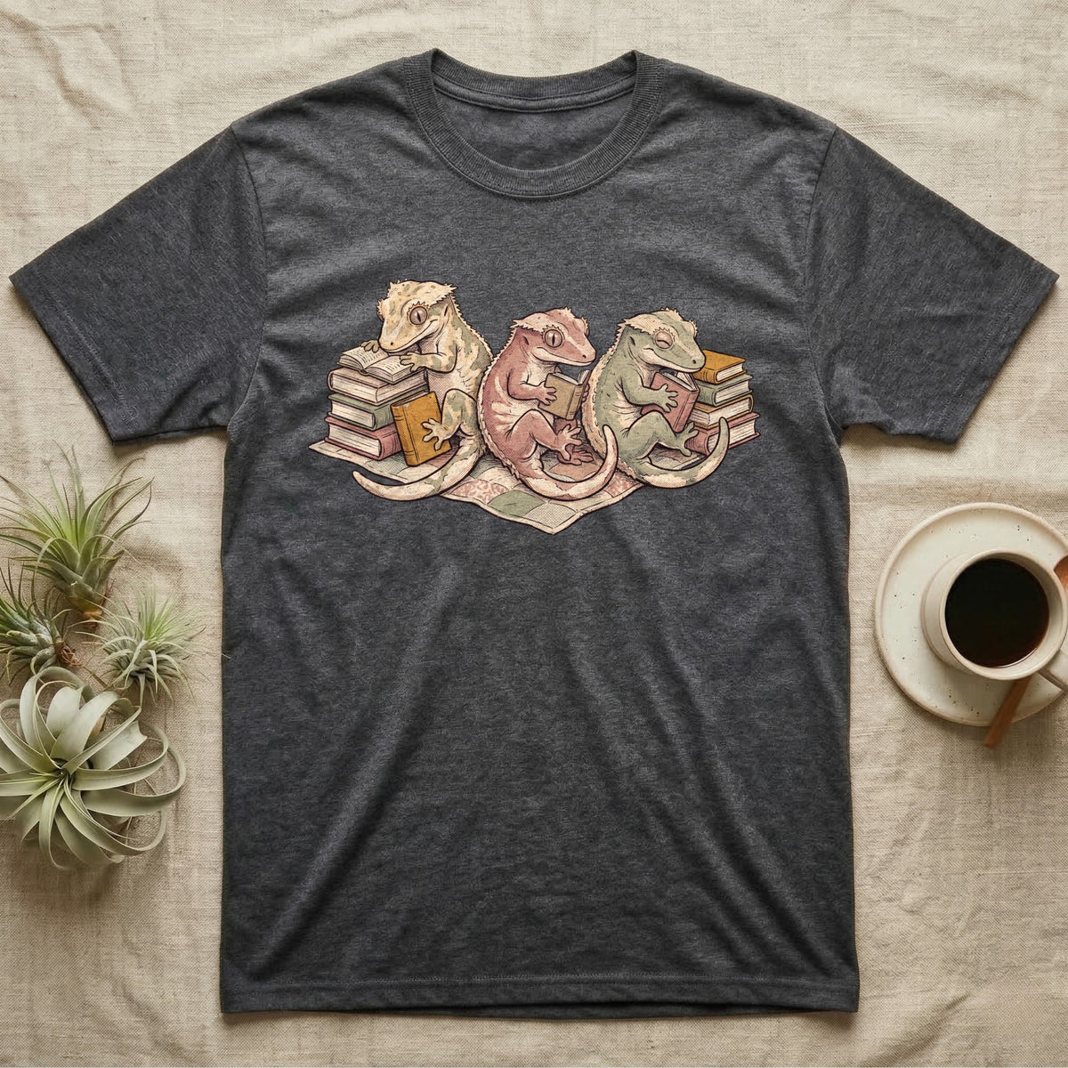 Gecko Book Club T-Shirt in Dark Heather — Warm storybook illustration of three crested geckos in a cozy reading row, each beside a tiny stack of books.
