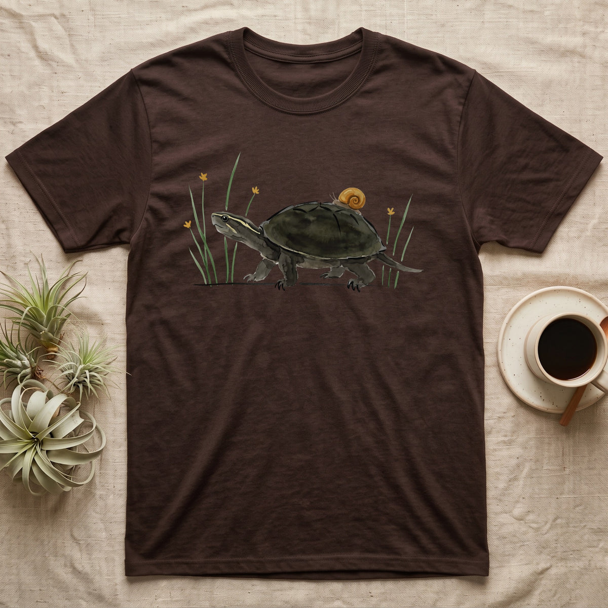 Musk Turtle Hitchhiker T-Shirt in Dark Chocolate — Watercolour musk turtle walking left with a ramshorn snail on its shell, beside slender grass blades with tiny amber flowers.