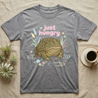 African Bullfrog Baby T-Shirt in Sport Grey — Pastel African bullfrog eyeing a tiny bug among reeds and flowers, puffy pink bubble wordmark "just hungry" above.