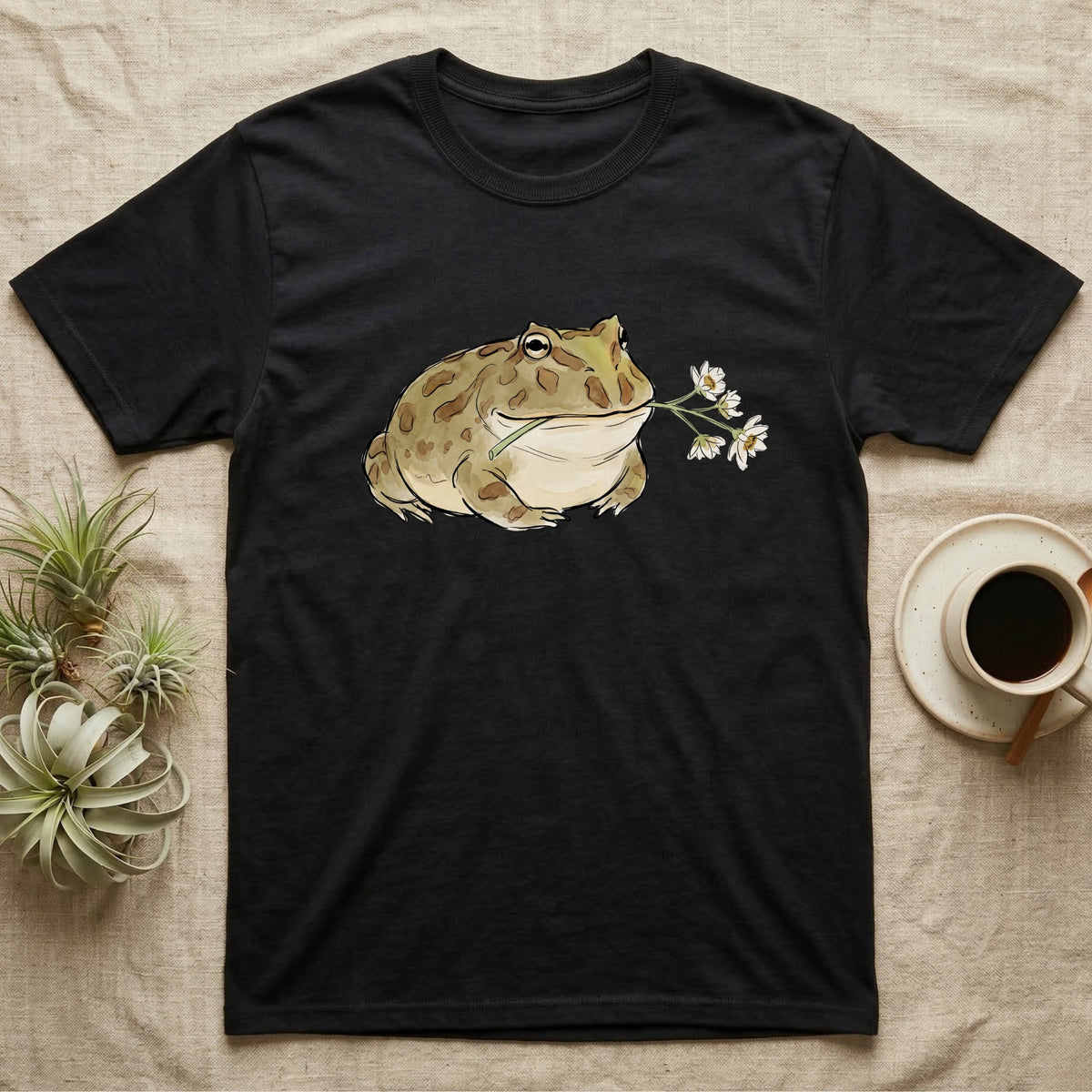 Pacman Frog Blossom T-Shirt in Black — Watercolor illustration of an olive-tan Pacman frog holding a water-lily blossom sprig, on a dark grey t-shirt.