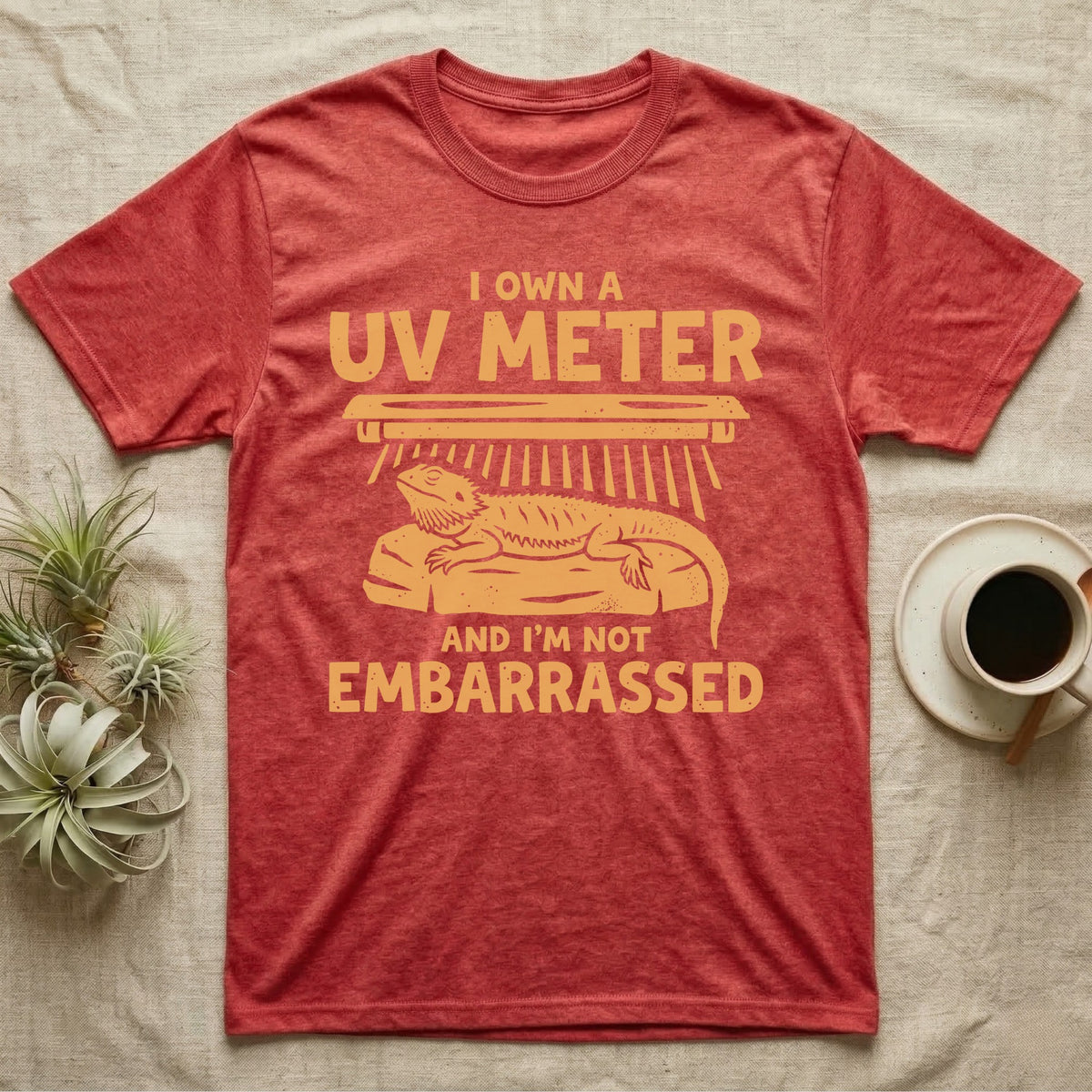 Reptile Keeper UV Meter T-Shirt in Heather Red — Linocut block-print badge of a UVB tube lamp above an empty basking rock, ringed by carved slogan lettering.