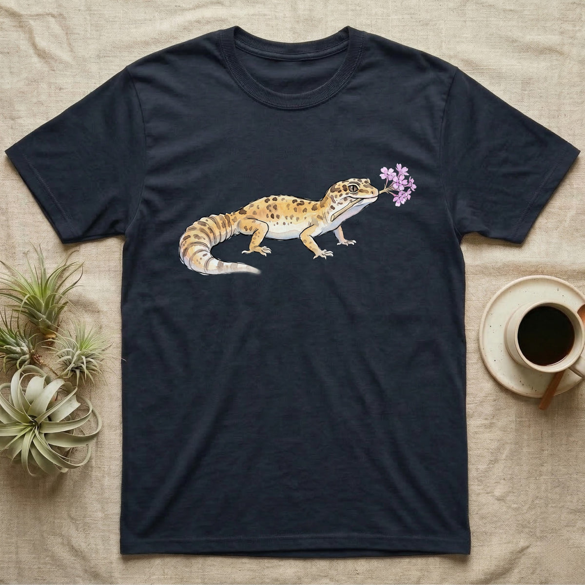 Leopard Gecko Blossom T-Shirt in Navy — Watercolor illustration of a sandy-gold spotted leopard gecko holding a sand verbena blossom sprig, on a dark grey t-shirt.
