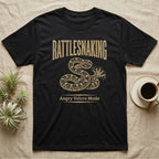 Coiled corn snake mid-tail-vibration in warm tan ink engraving beneath an arched wordmark reading RATTLESNAKING and the subtitle Angry Velcro Mode. — shown on a Black tee.