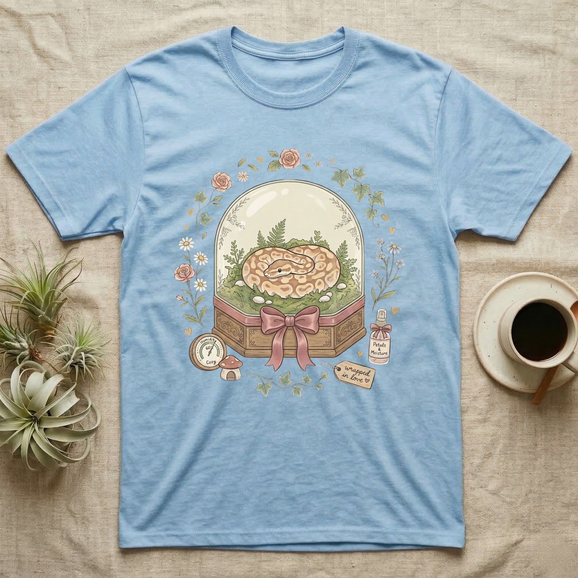 Ball python in a terrarium with flowers and "Wrapped in Love" text on a light blue t-shirt.