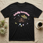 Tailless Whip Scorpion Baby T-Shirt in Black — Pastel tailless whip scorpion with long whip legs among leaves and blossoms, puffy pink wordmark "totally harmless" above.