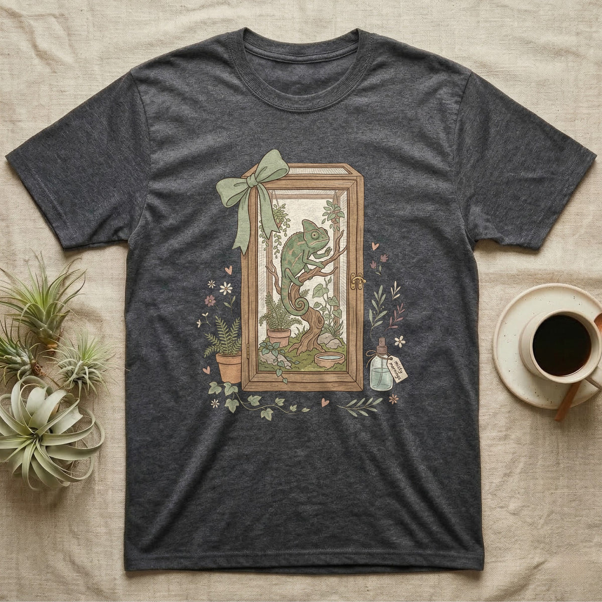 T-shirt graphic of a chameleon in a terrarium with plants and a green ribbon.