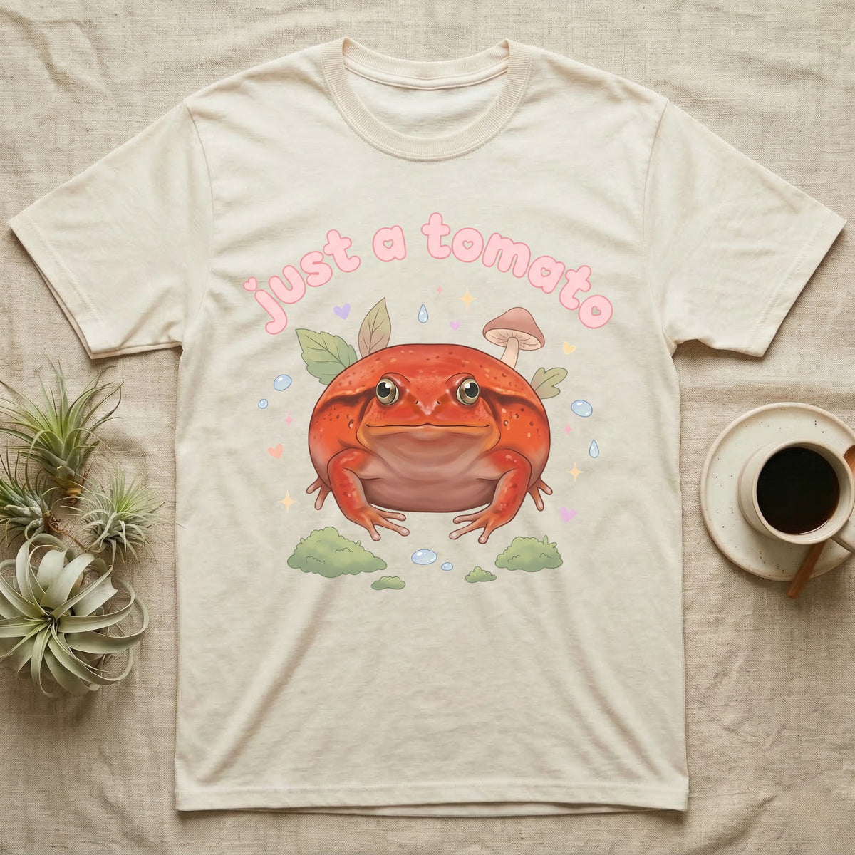Tomato Frog Softie T-Shirt in Natural — Round red-orange tomato frog facing forward under a puffy pastel "just a tomato" bubble wordmark with tiny hearts.