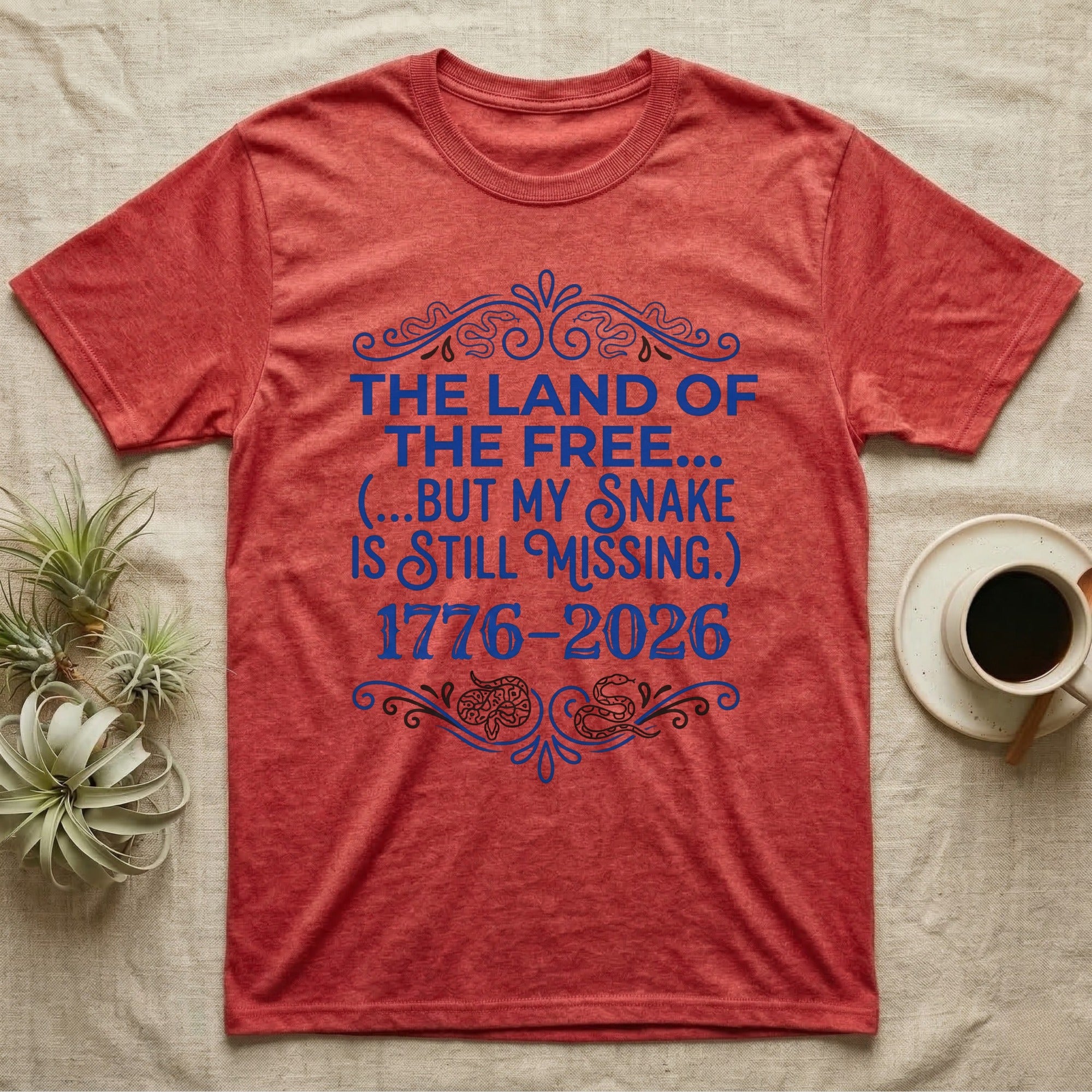 Corn Snake Still Missing Tee in Heather Red — Royal blue typographic tee reading The land of the free but my snake is still missing, with Victorian filigree and small coiled corn snakes.