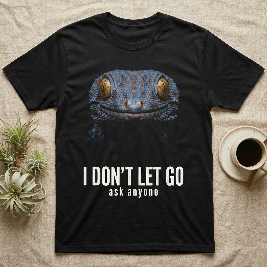 Deadpan close-up mugshot of a blue-grey tokay gecko on black, captioned I don't let go, ask anyone. — shown on a Black tee.