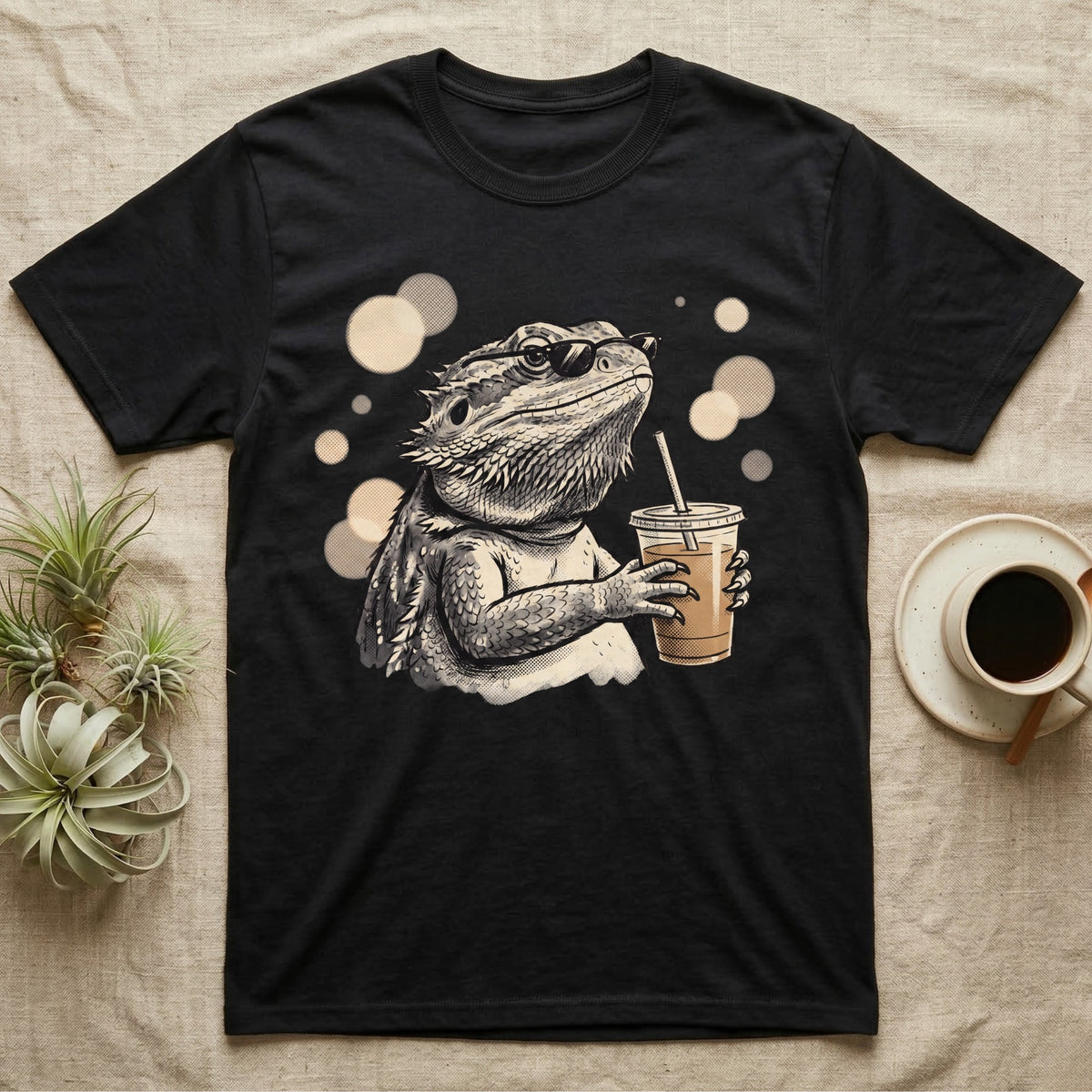 Cold Brew Bearded Dragon T-Shirt in Black — Flat near-photoreal illustration of a bearded dragon in tiny sunglasses holding a cold brew with warm bokeh.