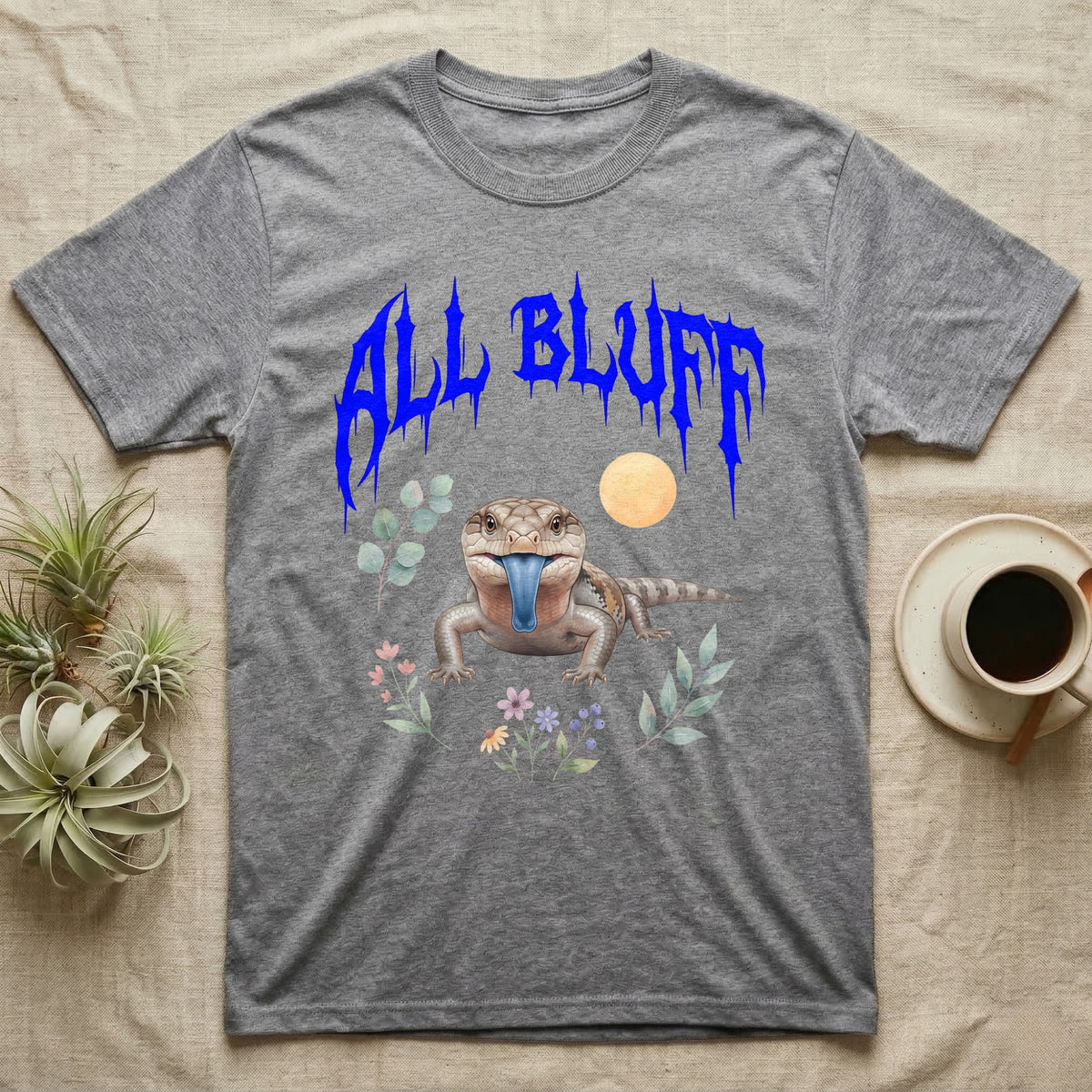 Northern Blue-Tongue Skink Feral T-Shirt in Sport Grey — Blue-tongue skink with its blue tongue out under a jagged cobalt-blue metal "ALL BLUFF" wordmark and pastel leaves.