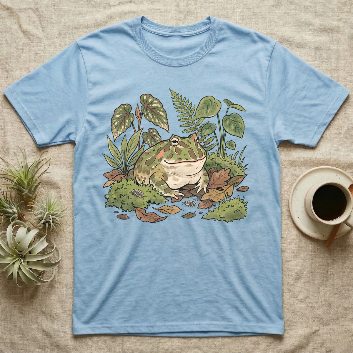Bioactive Pacman Frog T-Shirt in Light Blue — Warm botanical illustration of a content pacman frog half-buried in a lush bioactive habitat of moss and leaf litter.