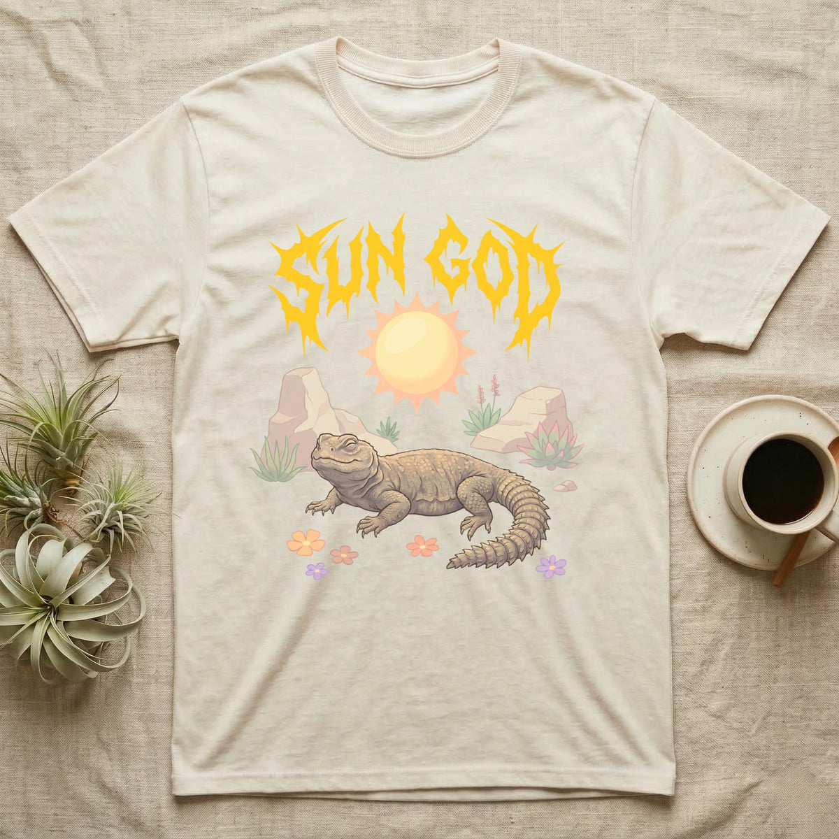 Moroccan Uromastyx Feral T-Shirt in Natural — Moroccan uromastyx basking under a big sun with a jagged solar-gold metal "SUN GOD" wordmark and pastel desert scene.