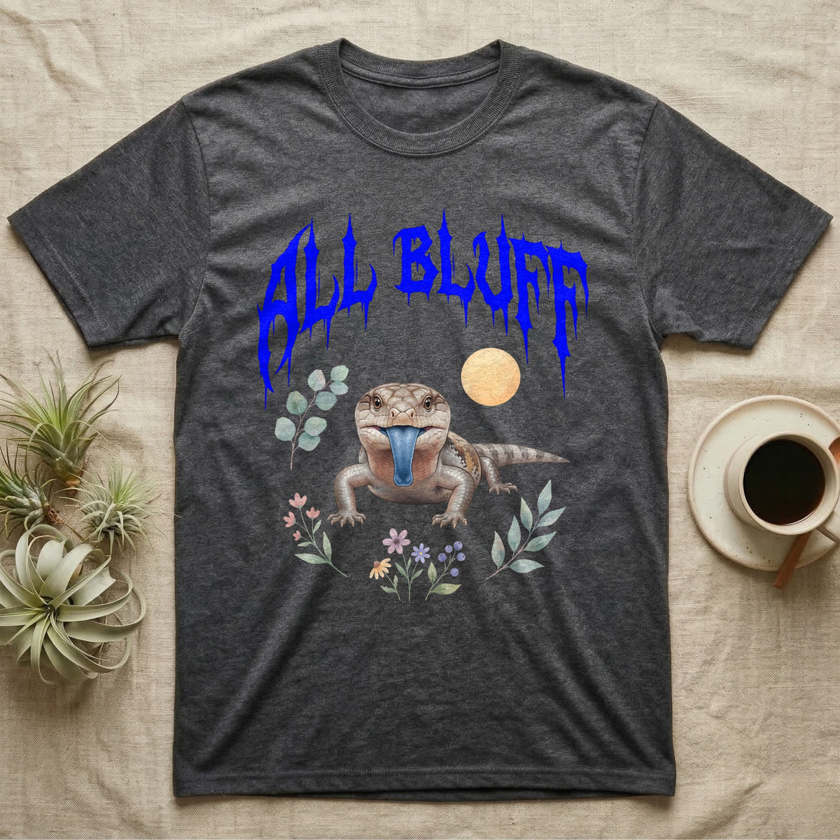 Northern Blue-Tongue Skink Feral T-Shirt in Dark Heather — Blue-tongue skink with its blue tongue out under a jagged cobalt-blue metal "ALL BLUFF" wordmark and pastel leaves.