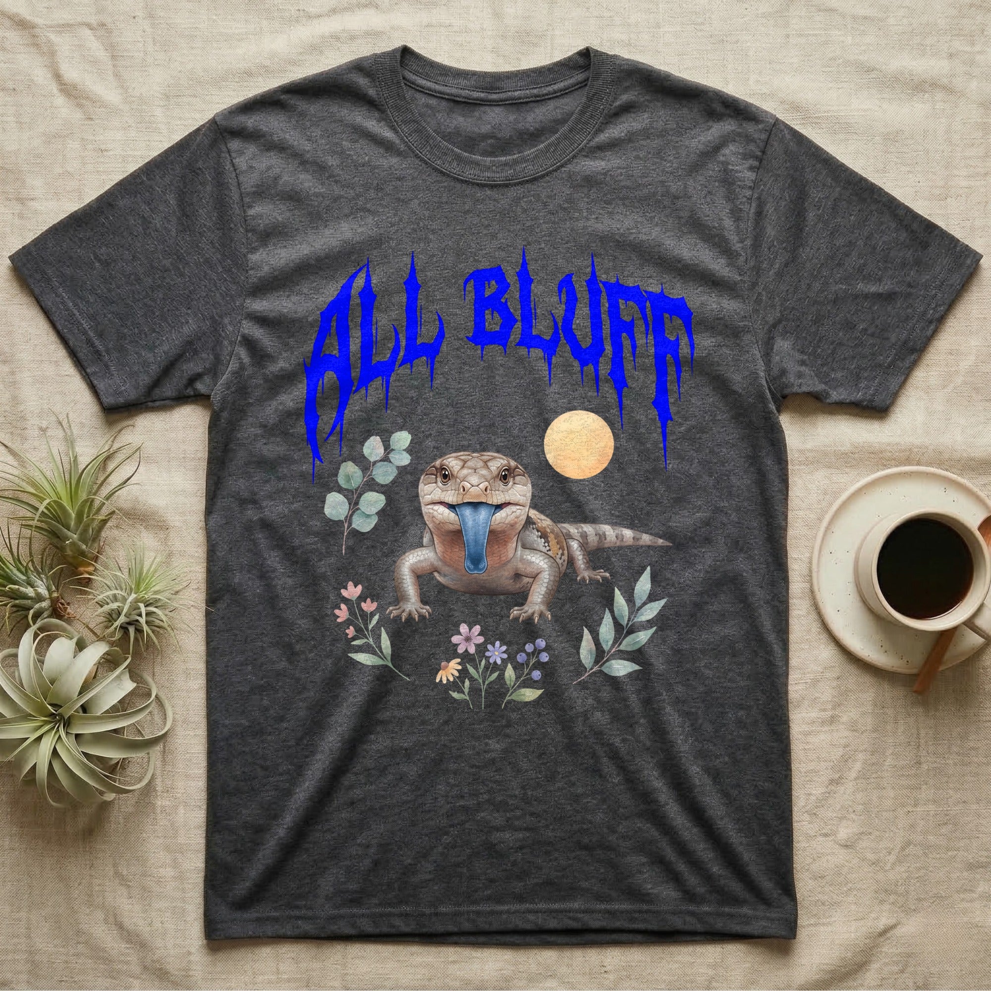 Northern Blue-Tongue Skink Feral T-Shirt in Dark Heather — Blue-tongue skink with its blue tongue out under a jagged cobalt-blue metal "ALL BLUFF" wordmark and pastel leaves.