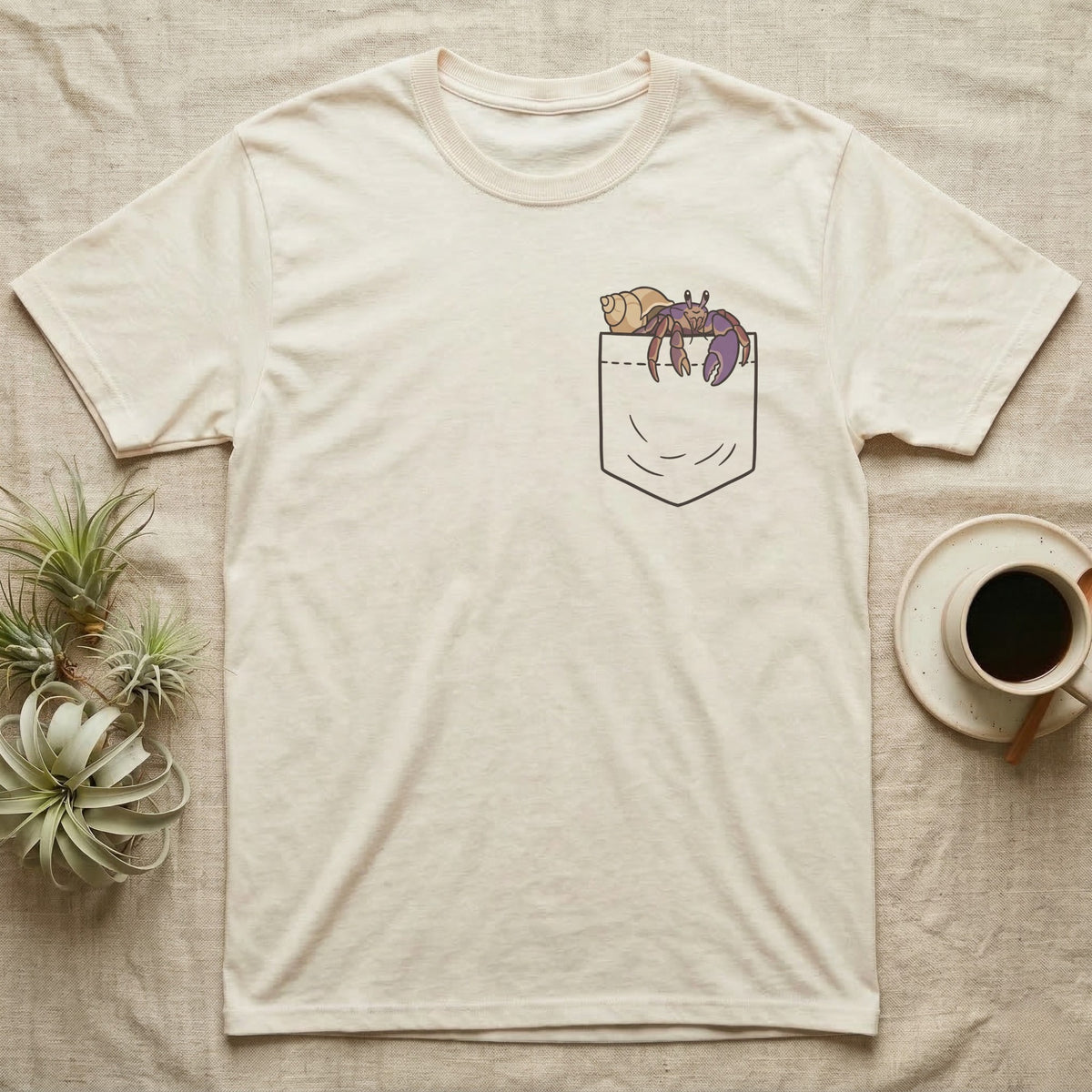Hermit Crab Pocket T-Shirt in Natural — Cartoon purple pincher hermit crab peeking out of a chest pocket, its empty shell on the pocket edge