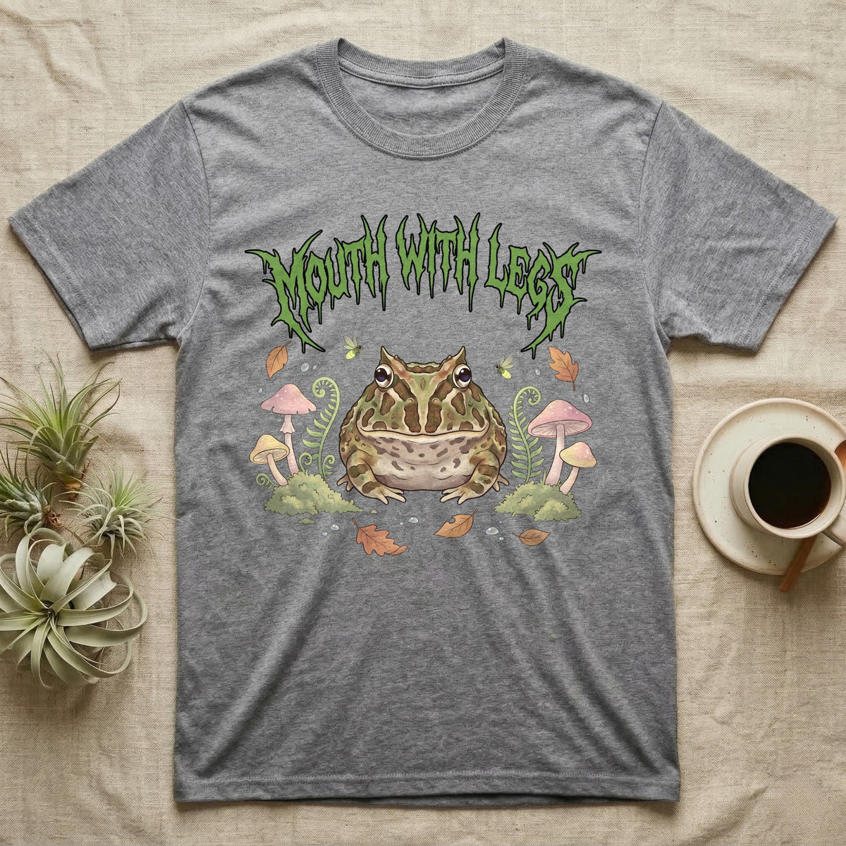 Pacman Frog Feral T-Shirt in Sport Grey — Round Pacman frog in a pastel forest-floor scene under a spiky leaf-green metal wordmark reading MOUTH WITH LEGS.