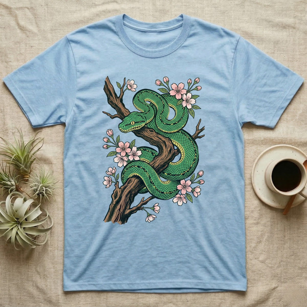 Green snake coiled on a tree branch with pink blossoms on a light blue t-shirt.