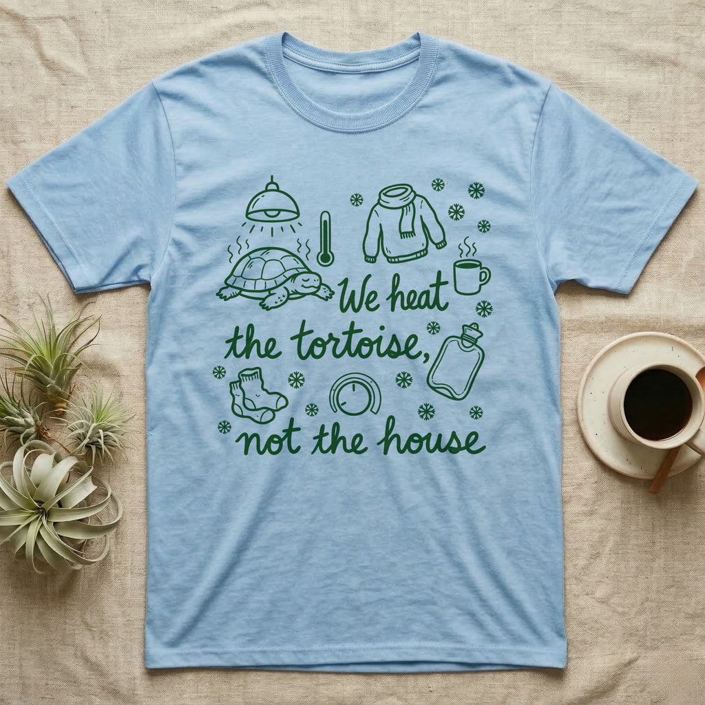 Russian Tortoise Winter T-Shirt in Light Blue — Green marker cursive reading We heat the tortoise, not the house: a tortoise basking under a heat lamp while socks, scarves and snowflakes fill the cold house