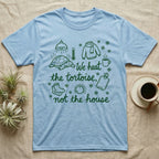 Russian Tortoise Winter T-Shirt in Light Blue — Green marker cursive reading We heat the tortoise, not the house: a tortoise basking under a heat lamp while socks, scarves and snowflakes fill the cold house