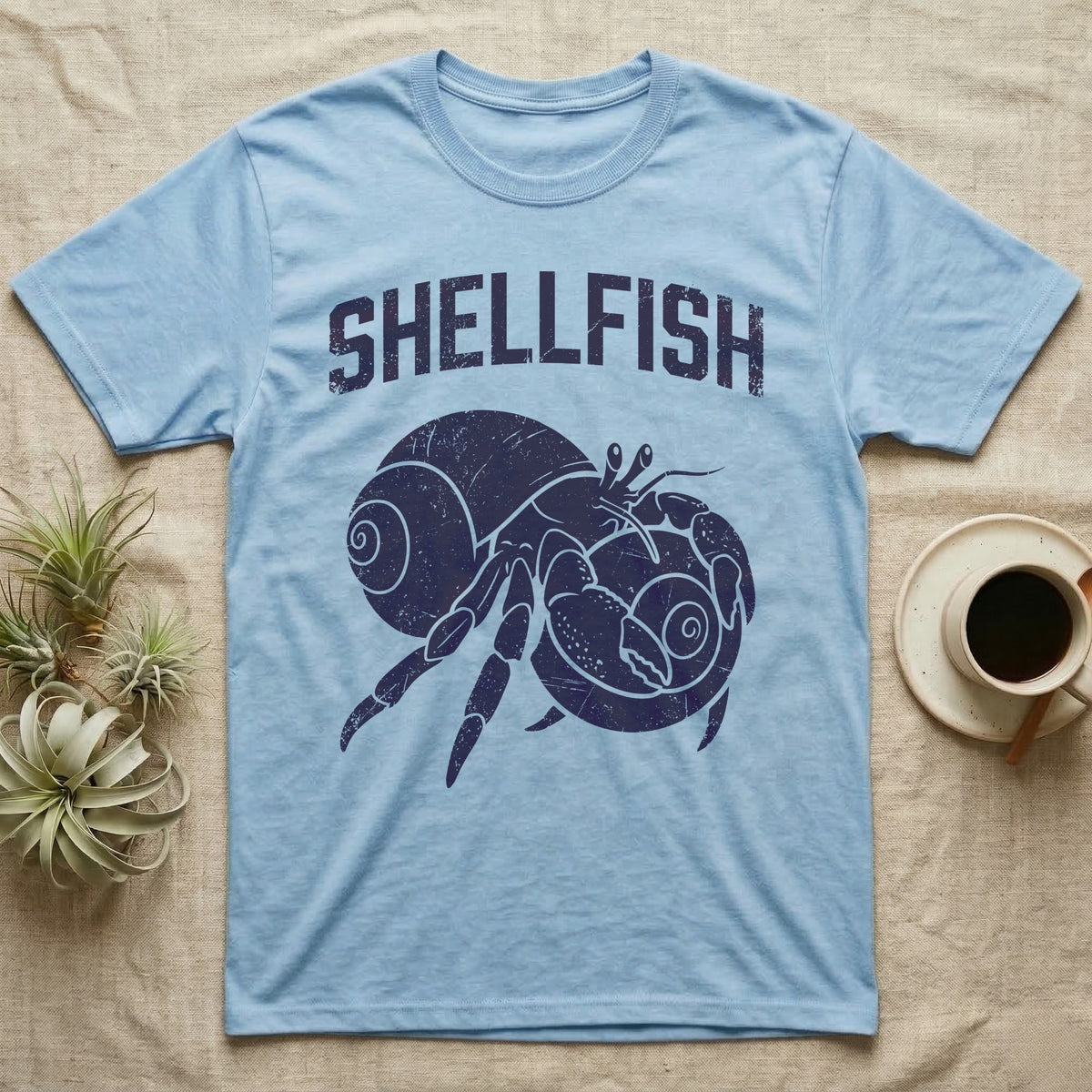 Hermit Crab Shellfish T-Shirt in Light Blue — Worn purple one-ink print of a hermit crab hugging a big spare shell to its chest under the word Shellfish