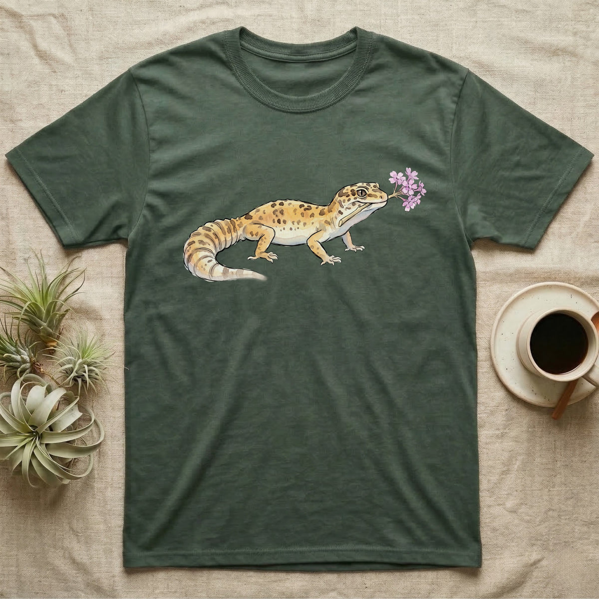 Leopard Gecko Blossom T-Shirt in Military Green — Watercolor illustration of a sandy-gold spotted leopard gecko holding a sand verbena blossom sprig, on a dark grey t-shirt.