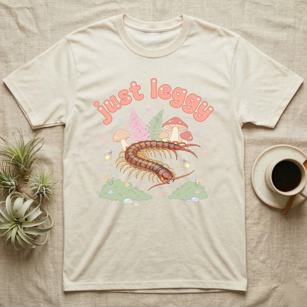 Vietnamese Centipede Baby T-Shirt in Natural — Many-legged Vietnamese centipede curving among pastel ferns and toadstools under puffy coral text "just leggy".