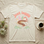 Vietnamese Centipede Baby T-Shirt in Natural — Many-legged Vietnamese centipede curving among pastel ferns and toadstools under puffy coral text "just leggy".