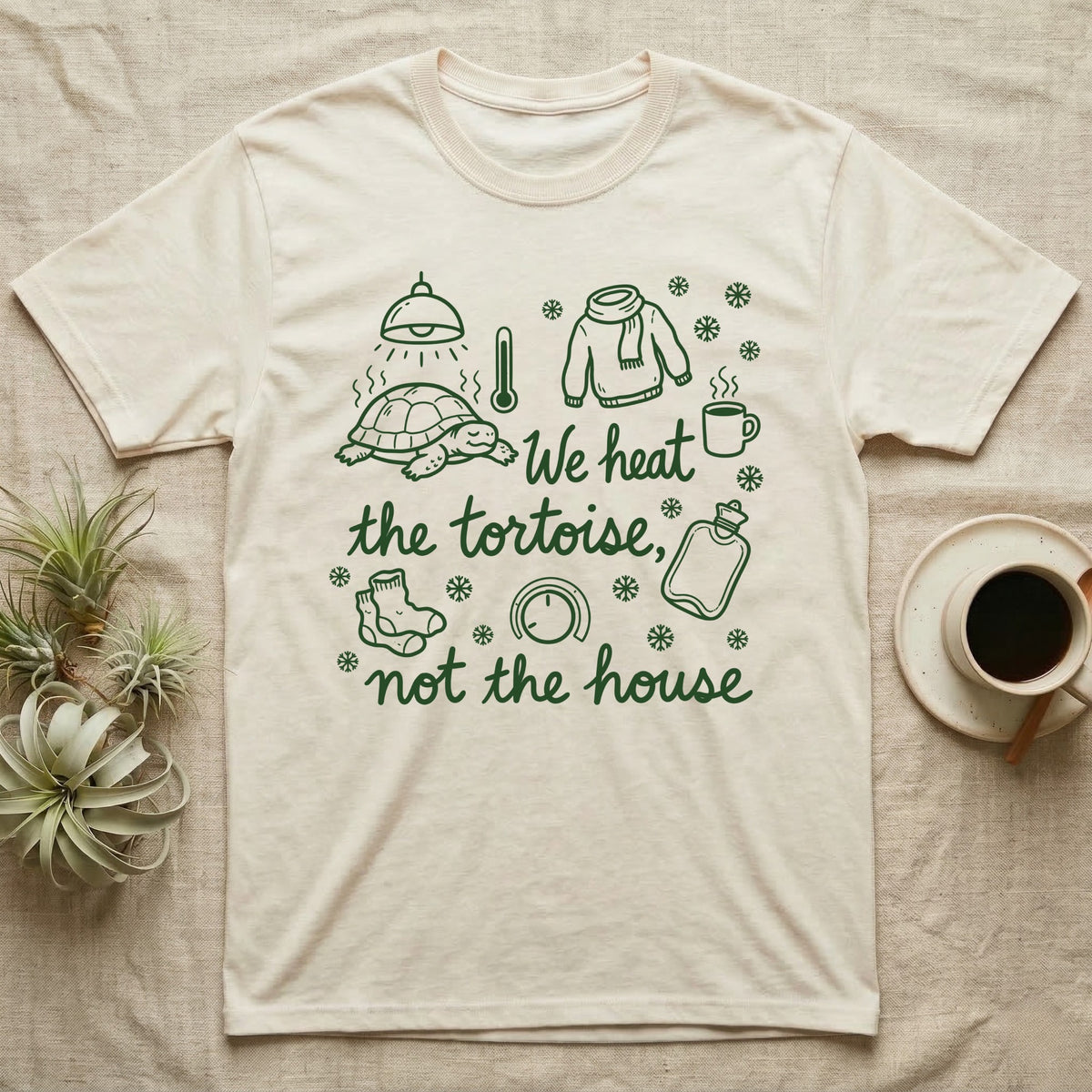 Russian Tortoise Winter T-Shirt in Natural — Green marker cursive reading We heat the tortoise, not the house: a tortoise basking under a heat lamp while socks, scarves and snowflakes fill the cold house