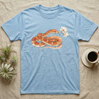Corn Snake Blossom T-Shirt in Light Blue — Watercolor illustration of an orange-and-cream corn snake holding a magnolia blossom sprig, on a dark grey t-shirt.