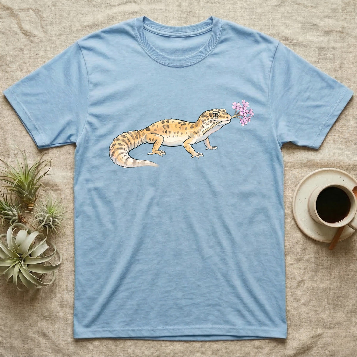 Leopard Gecko Blossom T-Shirt in Light Blue — Watercolor illustration of a sandy-gold spotted leopard gecko holding a sand verbena blossom sprig, on a dark grey t-shirt.
