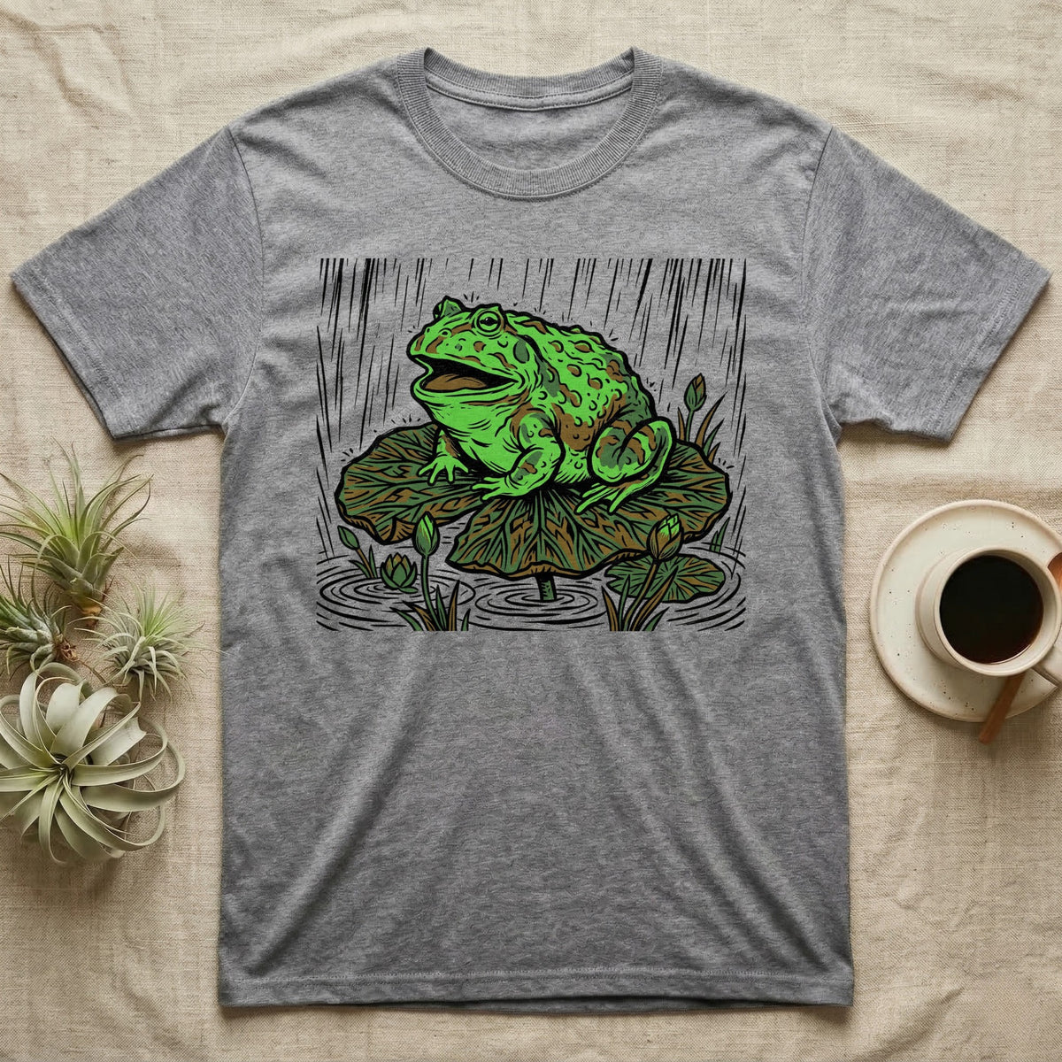 Gray t-shirt with illustration of a frog on a lily pad in the rain.