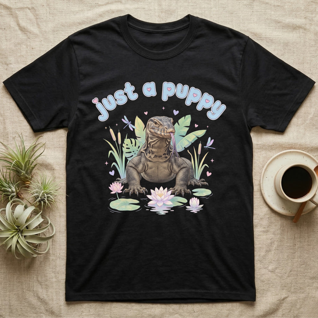 Asian Water Monitor Baby T-Shirt in Black — Asian water monitor with tongue out among pastel reeds and lotus under puffy blue bubble text "just a puppy".