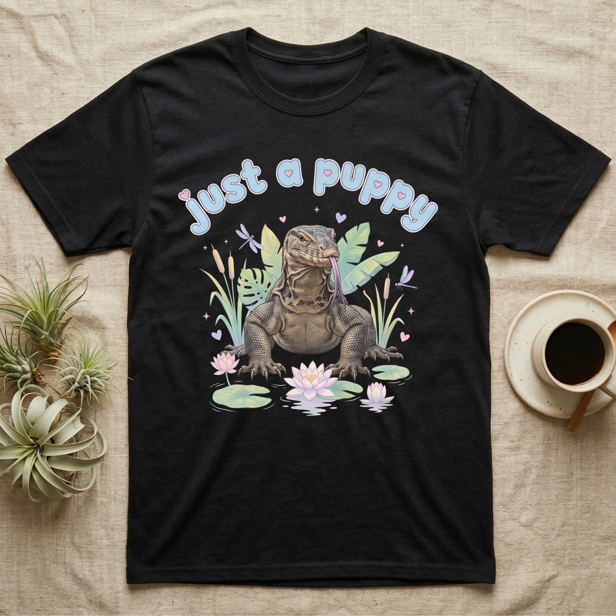 Asian Water Monitor Baby T-Shirt in Black — Asian water monitor with tongue out among pastel reeds and lotus under puffy blue bubble text "just a puppy".