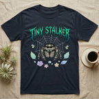 Regal Jumping Spider Feral T-Shirt in Navy — Fuzzy jumping spider with huge eyes in a pastel garden under a spiky iridescent-teal metal wordmark reading TINY STALKER.