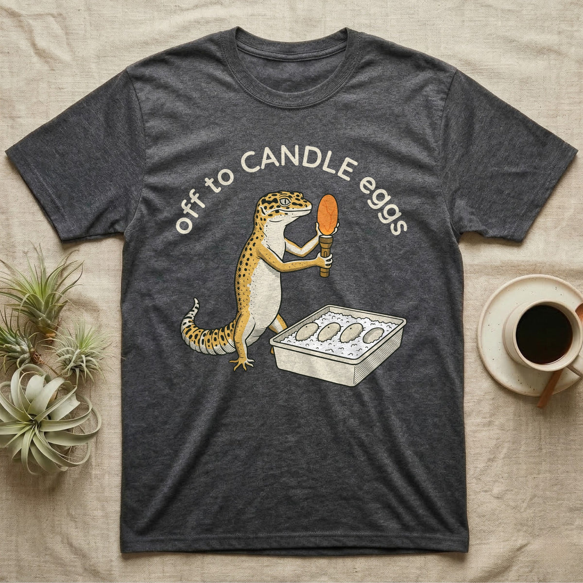 Leopard Gecko Candling T-Shirt in Dark Heather — Leopard gecko holding a torch under a glowing orange egg beside a tub of eggs, under the words off to CANDLE eggs