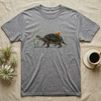 Musk Turtle Hitchhiker T-Shirt in Sport Grey — Watercolour musk turtle walking left with a ramshorn snail on its shell, beside slender grass blades with tiny amber flowers.