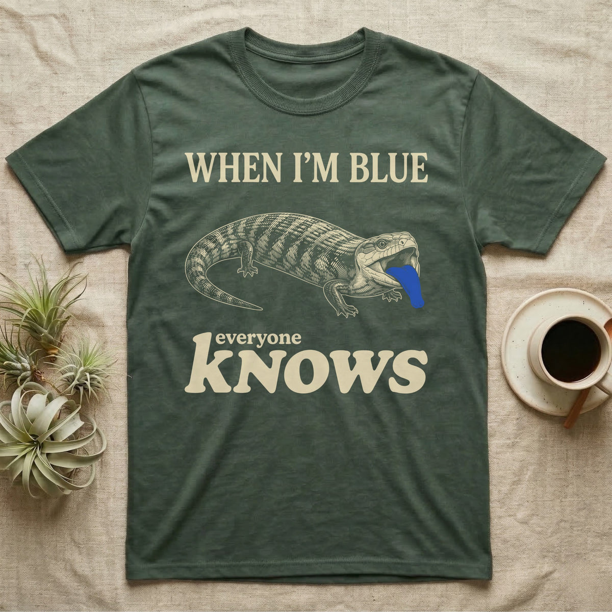 Blue-Tongue Skink Mood T-Shirt in Military Green — Cream engraved blue-tongued skink on black flashing a cobalt blue tongue under the words WHEN I'M BLUE everyone KNOWS