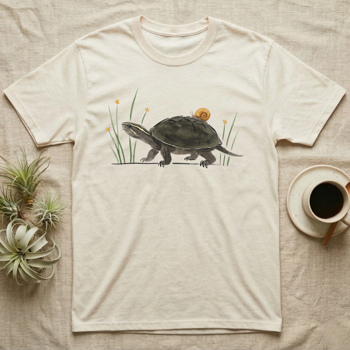 Musk Turtle Hitchhiker T-Shirt in Natural — Watercolour musk turtle walking left with a ramshorn snail on its shell, beside slender grass blades with tiny amber flowers.