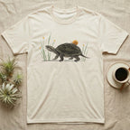 Musk Turtle Hitchhiker T-Shirt in Natural — Watercolour musk turtle walking left with a ramshorn snail on its shell, beside slender grass blades with tiny amber flowers.
