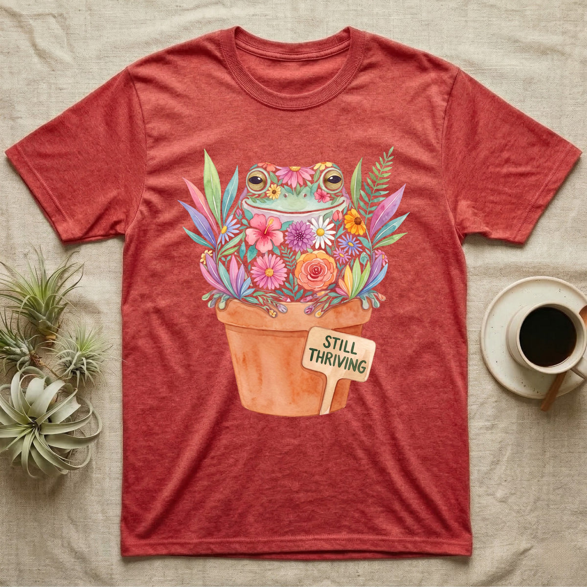 Tree Frog Thriving T-Shirt in Heather Red — A White's tree frog made of watercolour flowers, sitting in a terracotta pot with a plant label reading STILL THRIVING.