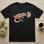 Crested Gecko Blossom T-Shirt in Black — Watercolor illustration of a rust-brown crested gecko holding a frangipani blossom sprig, on a dark grey t-shirt.