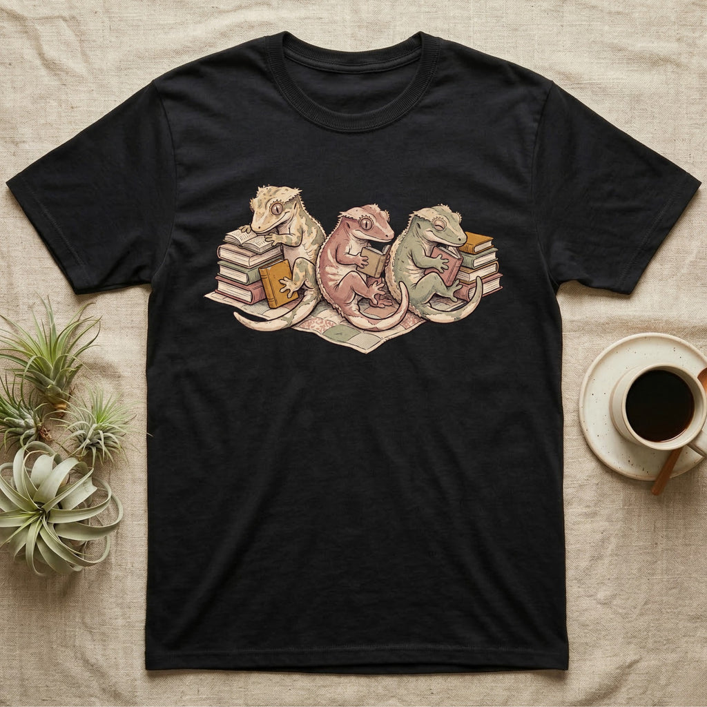Gecko Book Club T-Shirt in Black — Warm storybook illustration of three crested geckos in a cozy reading row, each beside a tiny stack of books.